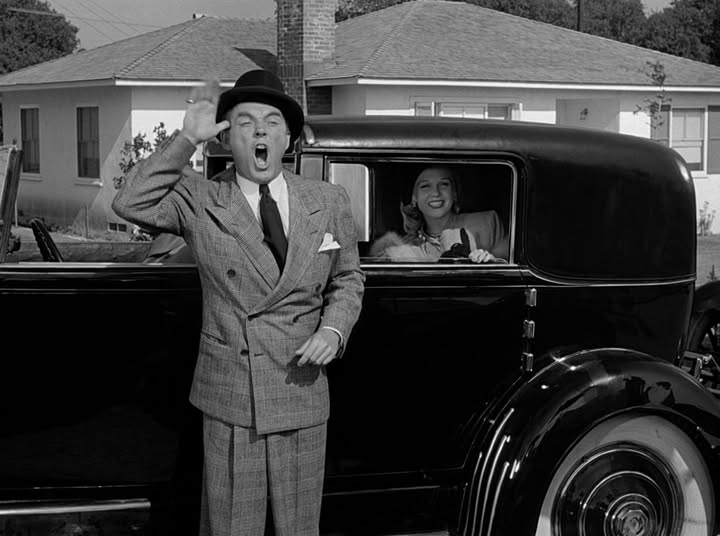man in a suit holds one hand to his temple to imitate a moose antler and his mouth is open wide