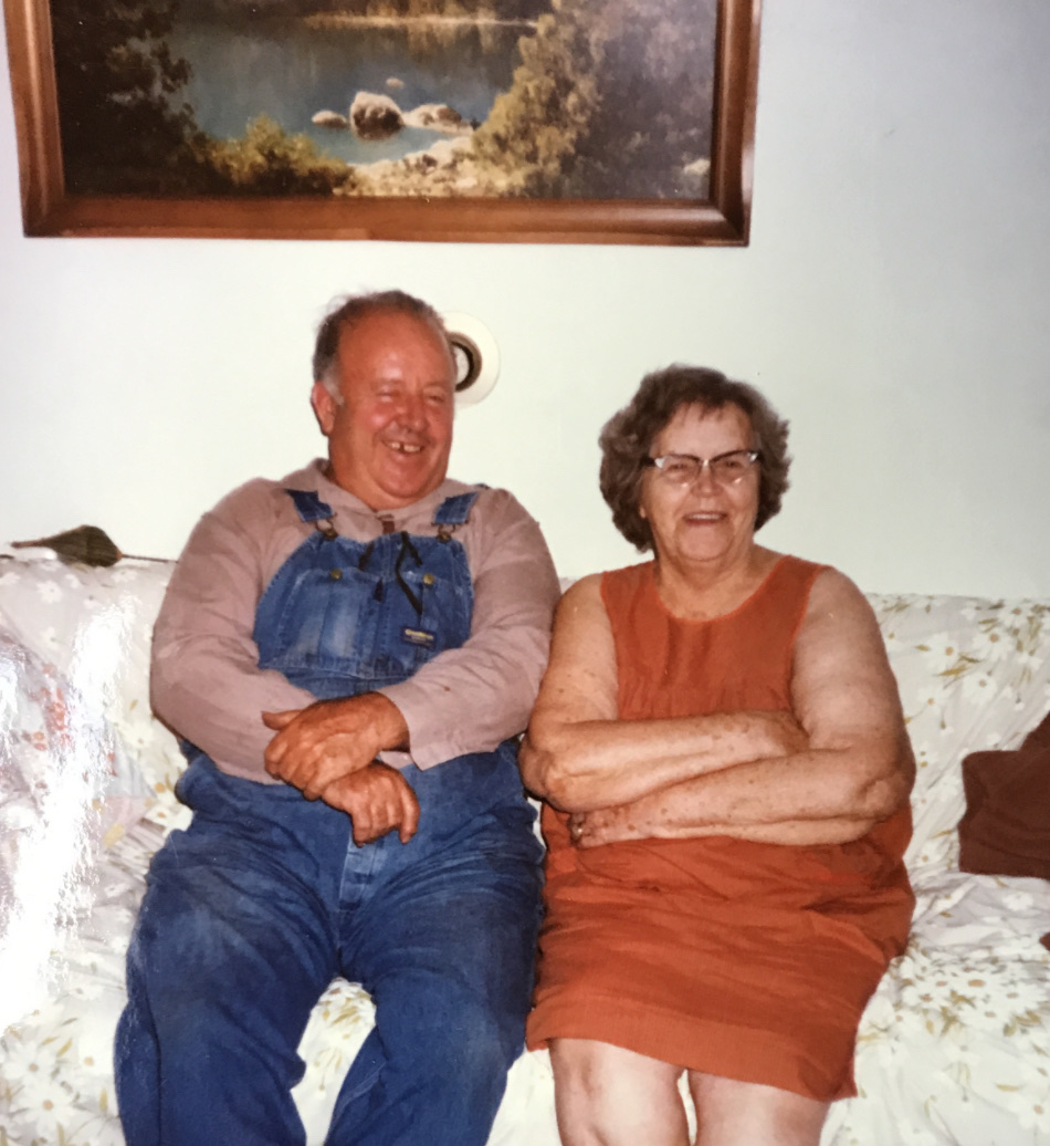 Man in bib overalls and woman in red dress sit on a couch smiling