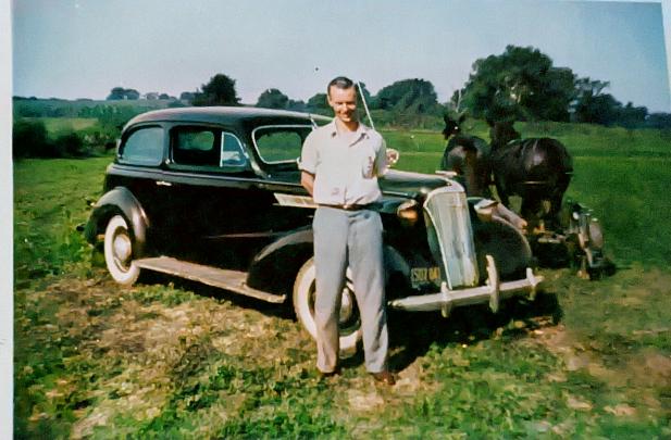 young man stands in front of 1940s style car parked in field next to a team of horses