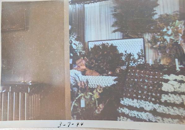 Man lies in casket with a flower wreath made in the shape of the US flag set in front