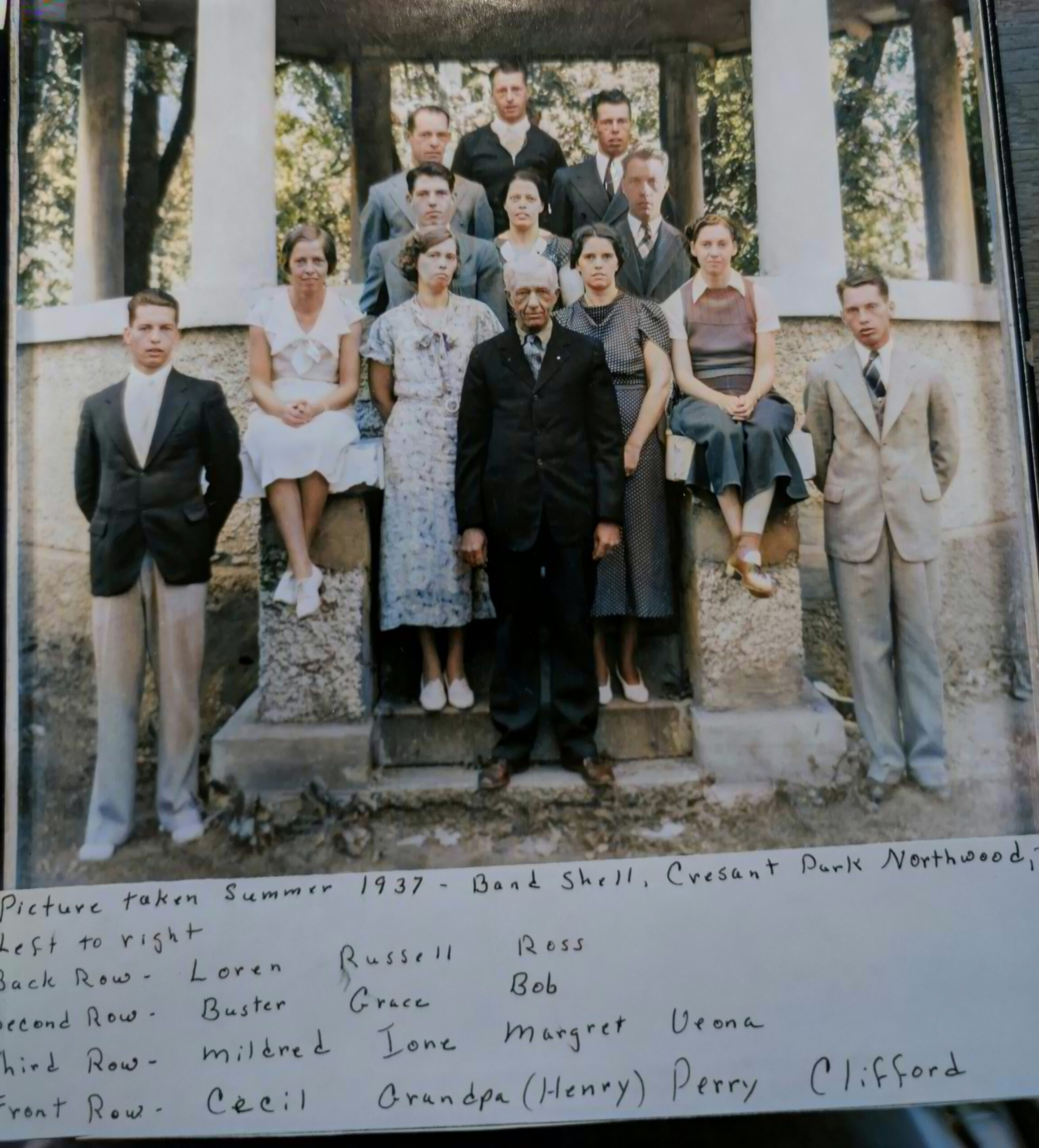 large family dressed in semi formal clothes of the 1930s 12 young adults stand behind an older man at an outdoor structure