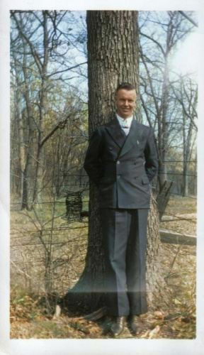 young man dressed in a double breasted suit and white shirt stands in front of a tree 