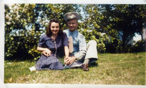 young caucasian couple, man in dress army uniform, girl in blue dress sitting on the grass, holding hands