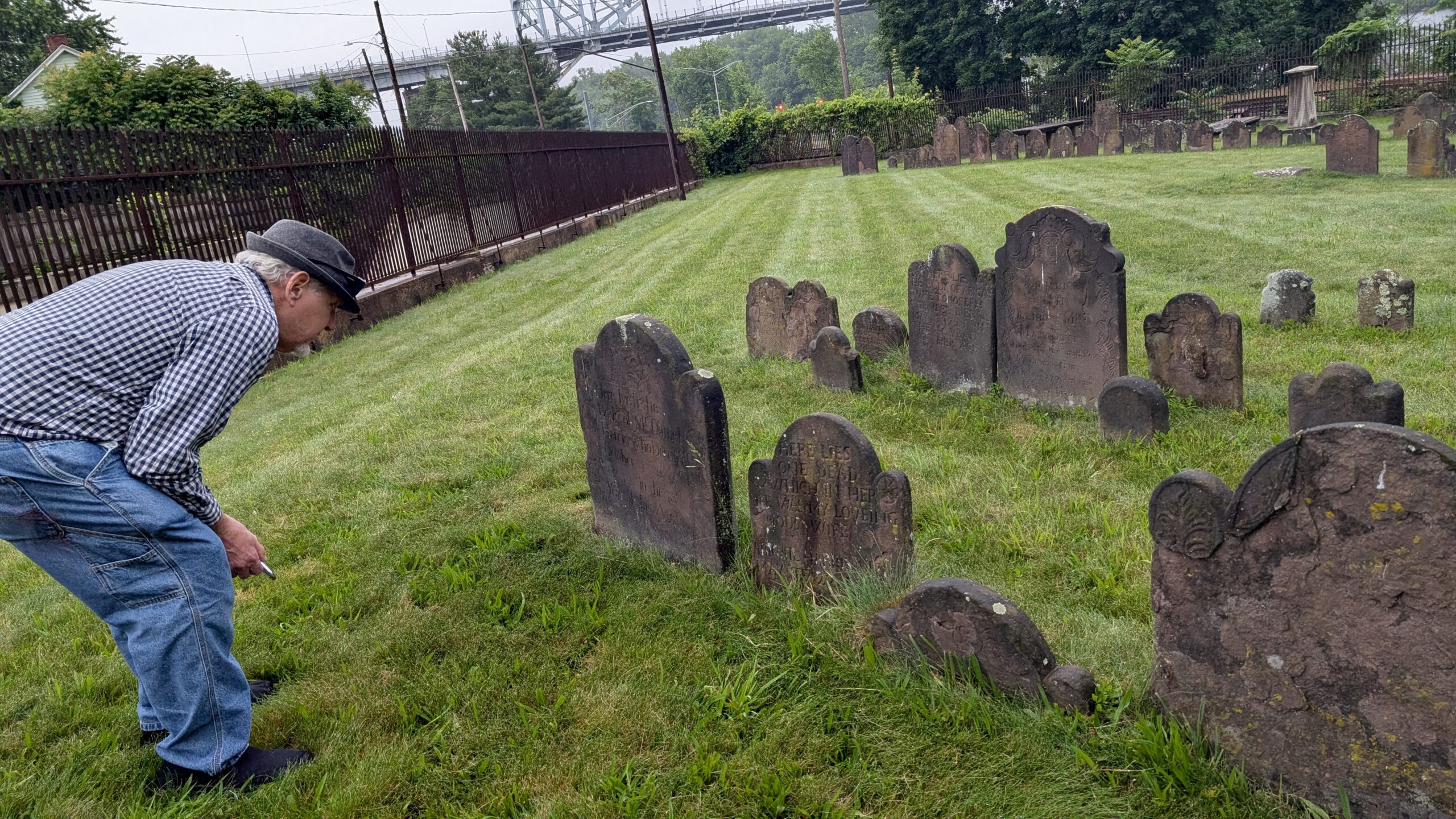 a white haired man bends down to examine a line of brownstone grave stones