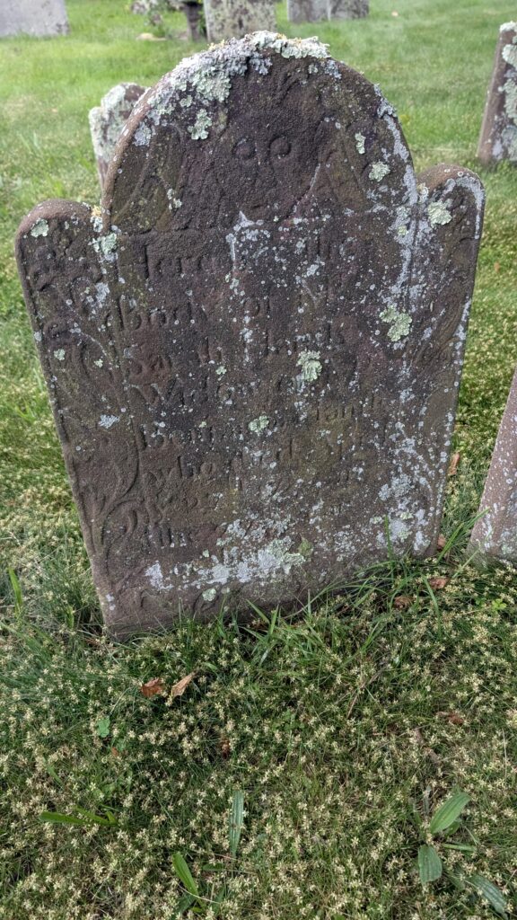 moss covered brownstone grave stone, a face engraved at the top, scroll work and the name Sarah Hands widow of Benjamin AGE 76 YEARS